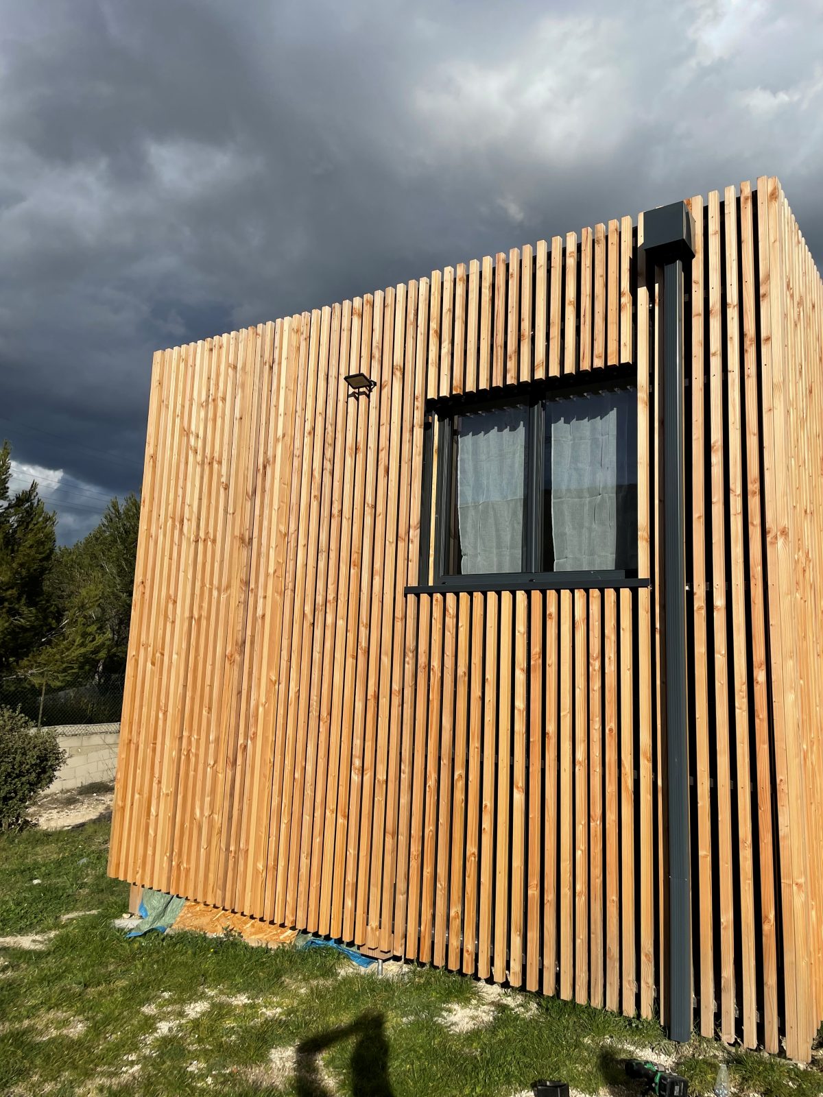 Façade bardage claire-voie et menuiserie noire sous ciel d'orage